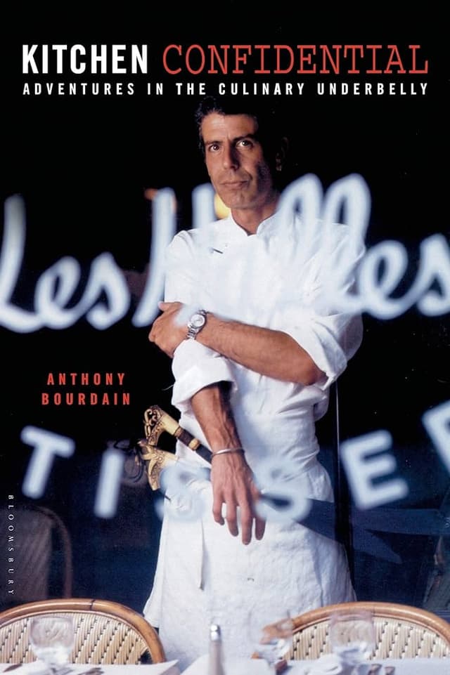Kitchen Confidential by Anthony Bourdain book cover which is a portrait of the late author in his chef's uniform standing behind a restaurant glass window and woven chairs